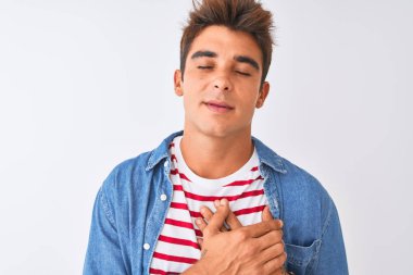 Genç yakışıklı adam kapalı gözleri ve yüzünde minnettar jest ile göğüs üzerinde elleri ile gülümseyerek izole beyaz arka plan üzerinde çizgili t-shirt ve denim gömlek giyiyor. Sağlık kavramı.