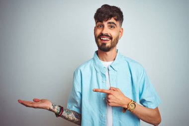 Dövme ile genç adam mavi gömlek ayakta izole beyaz arka plan hayran ve kamera için gülümseyen el ile sunmak ve parmak ile işaret.