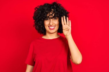 İzole kırmızı arka plan üzerinde rahat t-shirt giyen kıvırcık saçlı genç arap kadın gösteren ve emin ve mutlu gülümserken parmakları ile işaret.