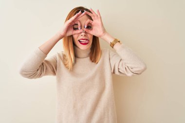 Kızıl saçlı kadın izole beyaz arka plan üzerinde duran zarif balıkçı yaka kazak giyen dürbün gibi ok jest yapıyor dil dışarı yapışıyor, gözleri parmaklar bakarak. Çılgın ifade.