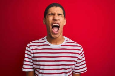 Genç yakışıklı Arap adam izole kırmızı arka plan üzerinde çizgili t-shirt giyen kızgın ve deli çığlık sinirli ve öfkeli, öfke ile bağırarak. Öfke ve agresif kavram.
