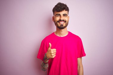 Dövme ile genç adam makyaj el ile jest kadar mutlu başparmak yapıyor izole pembe arka planda ayakta t-shirt. Başarı göstererek kameraya bakarak ifadeyi onaylama.