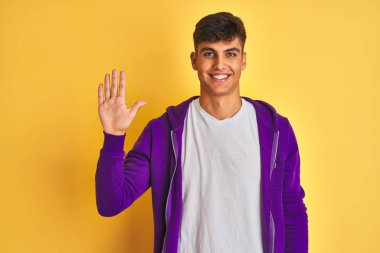 Genç hintli adam izole sarı arka plan üzerinde duran mor sweatshirt giyen gösteren ve emin ve mutlu gülümserken parmakları ile beş işaret.