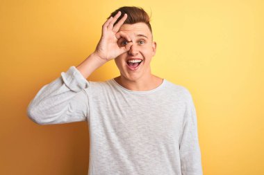 Beyaz rahat t-shirt giyen genç yakışıklı adam izole sarı arka plan üzerinde duran ok jest yaparken şaşırmış yüz ile şok, parmaklardan bakarak göz. İnanmayan ifade.