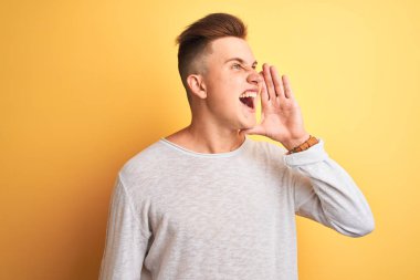 Genç yakışıklı adam beyaz rahat t-shirt giyerek izole sarı arka plan üzerinde ayakta bağırarak ve ağız da el ile yan yüksek sesle bağırıyor. İletişim kavramı.