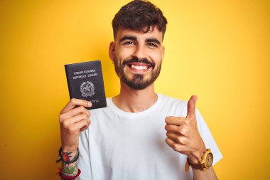 Genç adam dövme ile izole sarı arka plan üzerinde Italya Italyan pasaport giyen büyük bir gülümseme ile ok işareti, başparmak kadar parmak, mükemmel işareti yapıyor mutlu