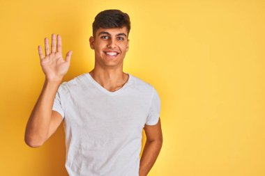 Beyaz tişört giyen genç hintli adam izole sarı arka plan üzerinde duran merhaba mutlu ve gülümseyen, samimi hoş geldin jesti söyleyerek Waiving