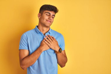 İzole sarı arka plan üzerinde duran mavi polo giyen genç hintli adam kapalı gözleri ve yüzünde minnettar jest ile göğüs üzerinde gülümseyerek. Sağlık kavramı.