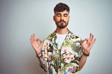Tatil üzerinde dövme ile adam izole beyaz arka plan üzerinde ayakta Yaz gömlek dikiliyor Relax ve gözleri ile gülümseyen meditasyon jest parmaklar ile kapalı. Yoga konsepti.