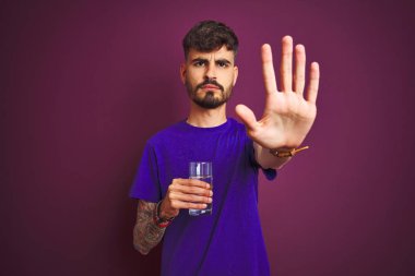 Açık el ile izole mor arka plan üzerinde ayakta su dövme içme bardağı ile genç adam ciddi ve emin ifade ile stop işareti yapıyor, savunma jest