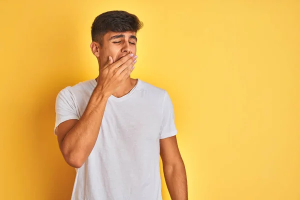 Young indian man wearing white t-shirt standing over isolated yellow background bored yawning tired covering mouth with hand. Restless and sleepiness.