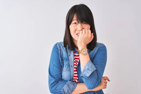 Chinese woman wearing denim shirt and red striped t-shirt over isolated white background looking stressed and nervous with hands on mouth biting nails. Anxiety problem.