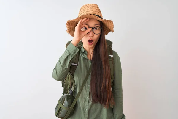 Chinese hiker woman wearing canteen hat glasses backpack over isolated white background doing ok gesture shocked with surprised face, eye looking through fingers. Unbelieving expression.