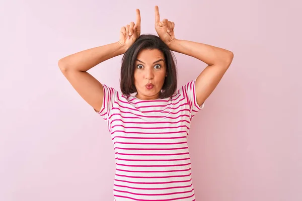 年轻的美女穿着条纹T恤站在孤立的粉红色背景做有趣的手势用手指在头上作为牛角 — 图库照片