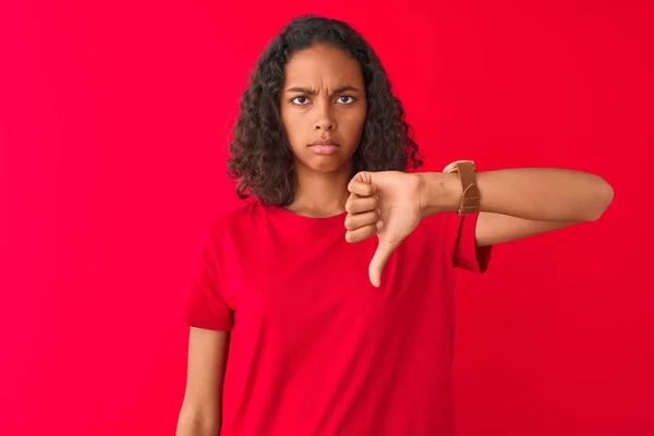 若いブラジル人女性が怒った顔で孤立した赤い背景の上に立っているTシャツを着て 親指を下にして嫌いを示す否定的なサイン 拒絶概念 — ストック写真