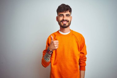 Dövme ile genç adam izole beyaz arka plan üzerinde ayakta turuncu kazak dikilerek el ile jest kadar mutlu başparmak yapıyor. Başarı gösteren kameraya bakarak ifadeyi onaylama.