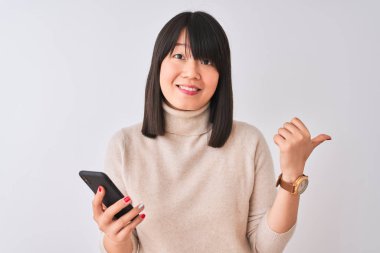 Genç güzel Çinli kadın izole beyaz arka plan üzerinde ayakta akıllı telefon kullanarak gösteren ve mutlu yüz gülümseyen yan başparmak ile gösteren