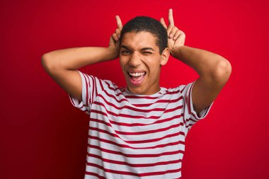Genç yakışıklı arap adam izole kırmızı arka plan üzerinde çizgili t-shirt giyen tavşan kulakları gibi baş parmakları ile komik ve deli poz, neşeli gülümseyen