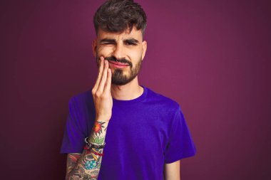 Dövme ile genç adam diş ağrısı ya da diş hastalıkları nedeniyle ağrılı ifade ile el ile izole mor arka plan dokunmadan ağız üzerinde t-shirt ayakta giyiyor. Dişçi konsepti.