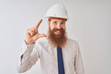 Genç Redhead İrlandalı mimar adam üzerinde güvenlik kask giyiyor izole beyaz arka plan gülümseyen ve emin işaret el ile küçük boyut işareti yapıyor parmaklar görünümlü ve kamera. Ölçü kavramı.