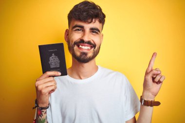 Dövme ile genç adam izole sarı arka plan üzerinde Kanada Kanada pasaport giyen çok mutlu yan el ve parmak ile işaret