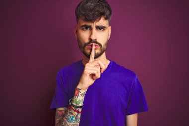 Dövme ile genç adam dudaklar üzerinde parmak ile sessiz olmak isteyen izole mor arka plan üzerinde ayakta t-shirt giyiyor. Sessizlik ve gizli konsept.