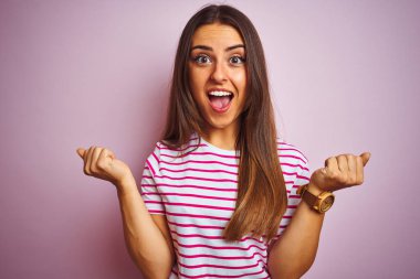 İzole pembe arka plan üzerinde ayakta çizgili t-shirt giyen genç güzel kadın şaşırttı ve silah kaldırdı ve açık gözleri ile başarı için hayran kutluyor. Kazanan kavramı.
