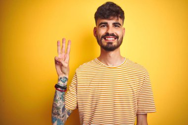 Dövme ile genç adam izole sarı arka plan üzerinde duran çizgili t-shirt ayakta giyen ve parmak sayısı üç ile işaret kadar emin ve mutlu gülümseyen.