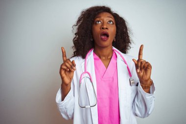 Izole beyaz arka plan üzerinde pembe stetoskop giyen Afrikalı Amerikalı doktor kadın şaşırttı ve yukarı bakarak ve parmakları ile işaret ve kolları kaldırdı şaşırttı.