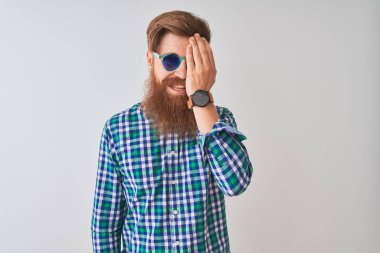 Rahat gömlek ve güneş gözlüğü giyen genç Redhead İrlandalı adam el ile bir göz kapsayan izole beyaz arka plan üzerinde, yüz ve sürpriz duygu kendine güvenen gülümseme.