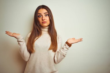 Genç güzel kadın izole beyaz arka plan clueless üzerinde kış balıkçı yakalı kazak giyiyor ve kollar ve elleri ile karıştı ifade kaldırdı. Şüphe kavramı.