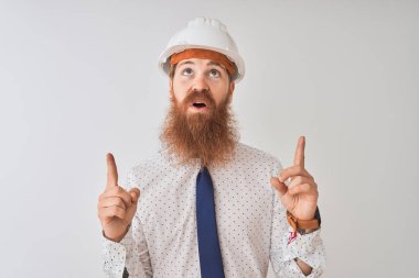 Genç Redhead İrlandalı mimar adam izole beyaz arka plan üzerinde güvenlik kask giyiyor şaşırdı ve görünümlü ve parmaklar ve yükseltilmiş kollar ile işaret şaşırttı.