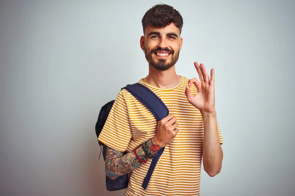 Young student man with tattoo wearing backpack standing over isolated white background doing ok sign with fingers, excellent symbol
