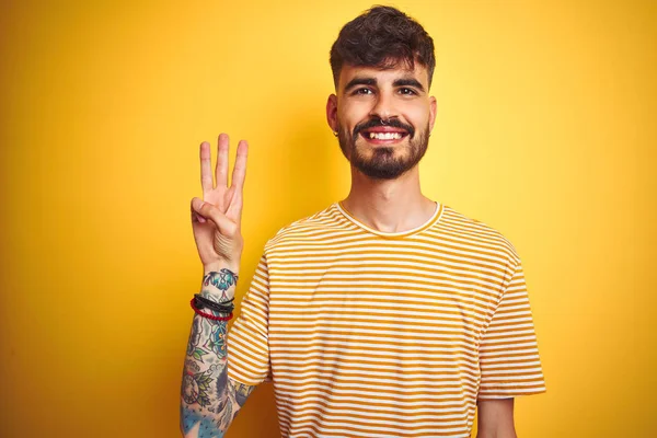 孤立した黄色の背景の上に立つストライプのTシャツを着た入れ墨をした若い男は 自信を持って幸せに微笑みながら 指番号3を示し 指を上げている — ストック写真