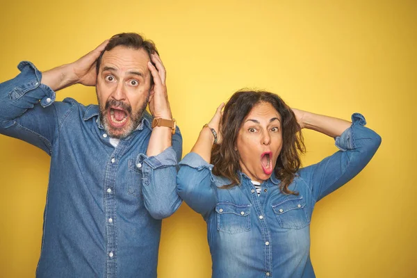 Beautiful middle age couple together standing over isolated yellow background Crazy and scared with hands on head, afraid and surprised of shock with open mouth