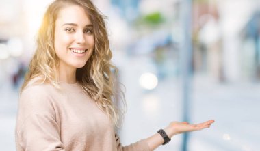 Açık el ile doğal gülümseyen girmek için davet izole arka plan üzerinde sweatershirt giyen genç güzel sarışın kadın