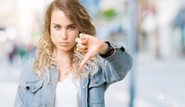 Mutsuz ve kızgın ret gösterilen ve başparmak aşağı hareketi ile negatif seyir izole arka plan üzerinde kot ceket giyen güzel genç sarışın kadın. Kötü ifade.