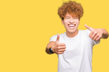 Afro saç ile genç yakışıklı adam rahat beyaz t-shirt el ile olumlu jest yapıyor onayladı giyen, thumbs yukarı gülümseyen ve başarı için mutlu. Kamera bakarak, kazanan jest.