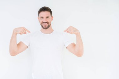Yüzünde gülümseme ile kendinden emin görünüyorsun, kendini gururlu ve mutlu parmakları ile işaret izole arka plan üzerinde rahat beyaz t-shirt giyen genç yakışıklı adam.