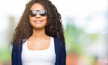 Kıvırcık saçlı giyen moda güneş gözlüğü ile mutlu ve serin güzel genç kızla yüzündeki gülümseme. Şanslı kişi.