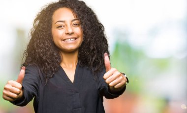 Kıvırcık saçlı onaylama zarif elbise giyen genç güzel kız el ile pozitif jest yapıyor gülümseyen ve başarı için mutlu yaşasın. Kamera, kazanan jest arıyorsunuz.