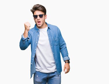 Genç yakışıklı adam izole arka plan üzerinde güneş gözlüğü giyiyor kızgın ve deli yükseltme yumruk sinirli ve öfkeli öfke ile bağırırken. Öfke ve agresif kavram.