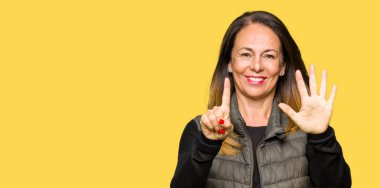 Güzel orta yaş kadın kış yelek gösteren giyen ve parmak numarası altı ile işaret ederken emin ve mutlu gülümseyen.