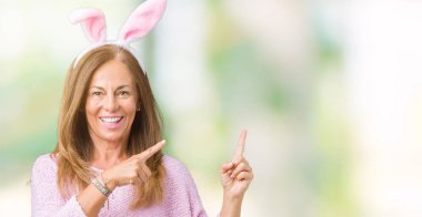 Orta yaş esmer kadın izole arka plan üzerinde Paskalya tavşan kulakları giyiyor gülümseyen ve iki el ve parmak tarafına işaret kamera bakıyor.