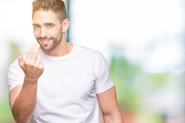 Açık havada arka planda beyaz t-shirt giyen yakışıklı adam beckoning el mutlu ve gülümseyen davet ile jest buraya geliyor