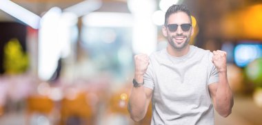 İzole arka plan üzerinde güneş gözlüğü takan genç yakışıklı adam gururlu çığlık ve zafer kutluyor ve başarı çok heyecanlı, tezahürat duygu