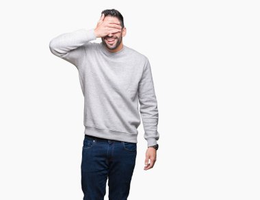 Genç yakışıklı adam gülümsüyor ve gözleri için sürpriz kapsayan yüz el ile gülüyor izole arka plan üzerinde kazak giyiyor. Kör kavramı.