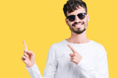 Yakışıklı delikanlı gülümseyen ve iki elleri ve parmakları ile tarafına işaret kamera bakarak izole arka plan üzerinde güneş gözlüğü takıyor.