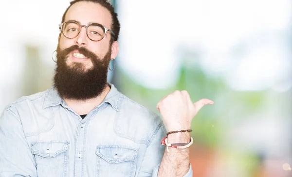 Young hipster man with long hair and beard wearing glasses smiling with happy face looking and pointing to the side with thumb up.