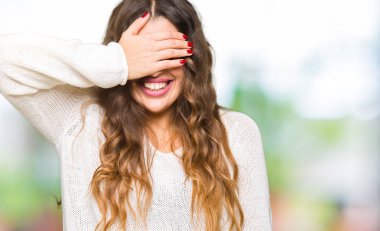 Beyaz kazak gülümsüyor ve gözleri sürpriz için kapsayan yüz el ile gülüyor giyen genç güzel kadın. Kör kavramı.